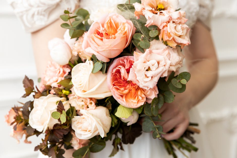 A person holding a floral bouquet arrangement featuring large, soft pink and peach roses, complemented by palest pink lisianthus and delicate peach-pink carnations, accented with fresh eucalyptus leav