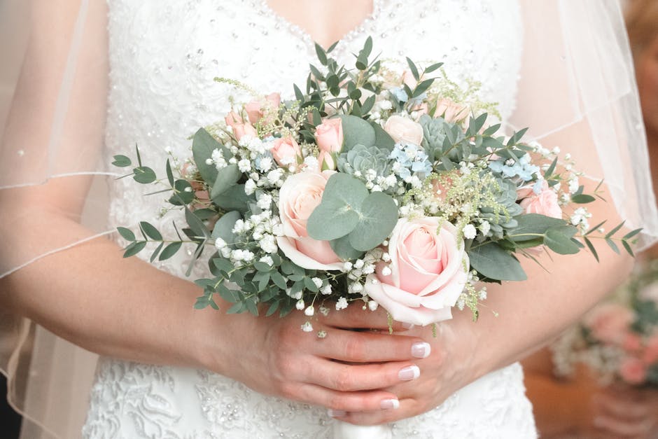 A bride is holding a floral bouquet arrangement featuring soft pink roses, white baby's breath, and green eucalyptus leaves, arranged in a loose, romantic style. The bouquet appears fresh, with velvet
