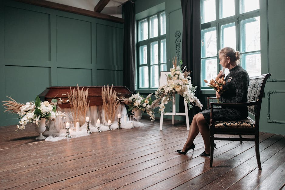 A woman in formal attire sits on a decorative upholstered chair in a softly lit room with green paneled walls and large windows, holding a small bouquet of flowers. In front of her, there is a memoria