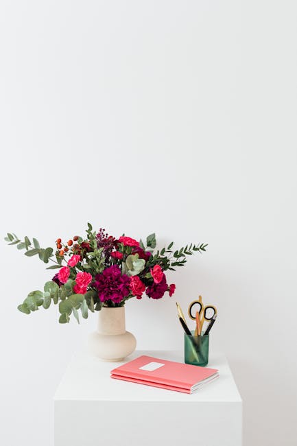 A floral arrangement featuring a bouquet of fresh flowers in a cream ceramic vase placed on a white table, with a mix of pink and deep red carnations, small clusters of red berries, and eucalyptus lea