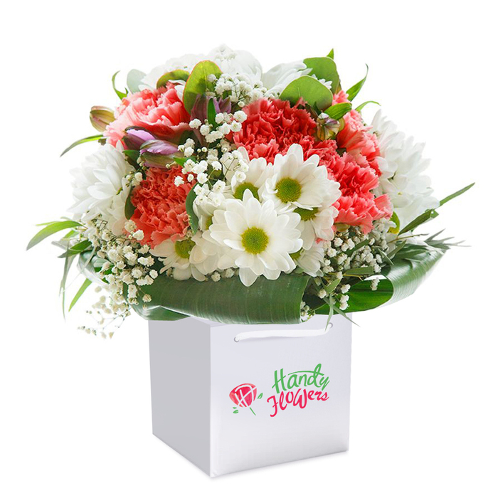 A lively, meticulously arranged bouquet evokes the best of Morden's floral tradition, captured as it stands upright in a chic white square gift bag. Each element is chosen for freshness and harmony: abundant coral and peach carnations ripple with intricate ruffled petals-soft to the eye and lush in form-while bright white daisies with charteuse-green centers punctuate the bouquet, lending it a cheerful, almost sunlit effect reminiscent of walks through Morden Hall Park on a warm afternoon. Baby's breath, in a scattering of airy white clusters, adds an elegant delicacy, playing off the dominant flowers' vibrancy. Closed purple-tinged buds nestle thoughtfully along one side-suggestive of the gentle unfolding of new beginnings and memories. The arrangement is brilliantly encircled by a generous collar of deep, glossy green leaves, arching outwards to cradle the blooms and provide a verdant, radiant backdrop. The sturdy white bag and its soft ribbon handles create an inviting, contemporary frame perfect for easy gifting or surprise same-day delivery across Morden and Wimbledon. Beautifully illuminated against a seamless white background, the bouquet's colours and softness are gently brought to life, presenting an ideal way to convey congratulations, gratitude, or loving sentiment to someone special. The artistry reflects not only floristry skill but also a sense of local connection and heartfelt care.