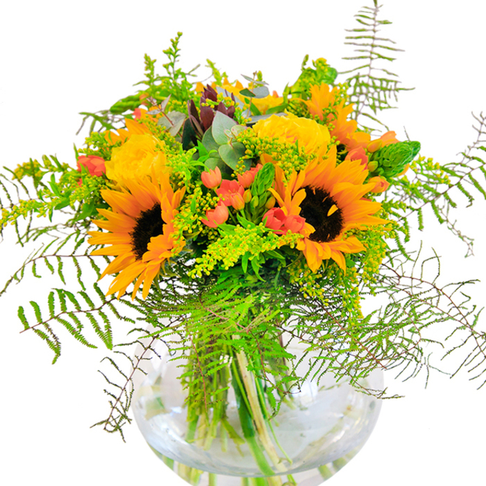 Bursting with radiant warmth, this floral arrangement brings a touch of Morden's garden-rich heritage indoors, presented in an elegant, bowl-shaped glass vase. Sunflowers command the eye, their golden petals and deep onyx centres echoing the vibrancy of summer afternoons spent in Morden Park. Interwoven among these, clusters of petite yellow filler blossoms evoke the wild hedgerows and meadows characteristic of the district. Delicate coral-orange accent flowers, cup-shaped and softly rounded, peek through lush greenery, their shade reminiscent of late sunset skies behind the Civic Centre. Lively fern fronds, arching and feathery, provide the bouquet with natural texture and movement, like the edges of an untamed woodland path, while sprigs of eucalyptus in calming blue-green tones introduce a contemporary twist. Pops of bright lime-green from the youngest foliage invigorate the display, creating seamless transitions between colours and textures. The clear vase, revealing the fresh green stems in sparkling water, underscores the bouquet's vitality-each bloom thoughtfully chosen by Morden Florist for artistry and freshness. With gentle, diffused light accentuating every vivid hue, the overall effect is uplifting and rejuvenating, suitable for birthday celebrations, friendly visits, or simply to brighten a Morden home with a dose of local floral elegance.